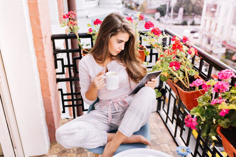 View from above pretty girl in pajama having breakfast on balcony in the morning in city. She holds a cup, reading on