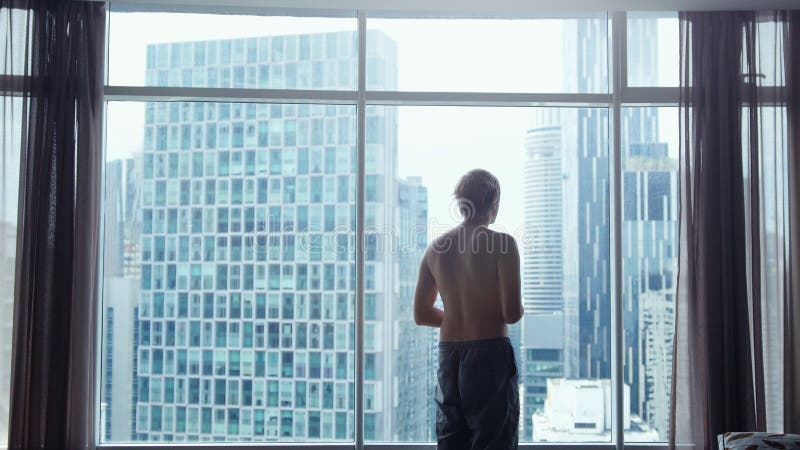 View from behind of young man in shorts looking out at view of the city. View from the window shows skyline with iconic