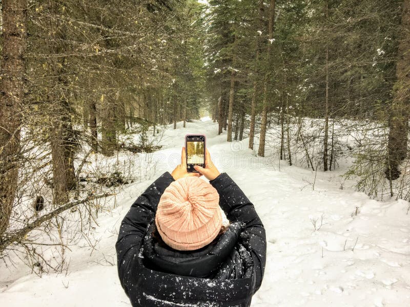 A view from behind of a young woman taking a photo on her phone while walking alone a snowy trail in the woods in nature