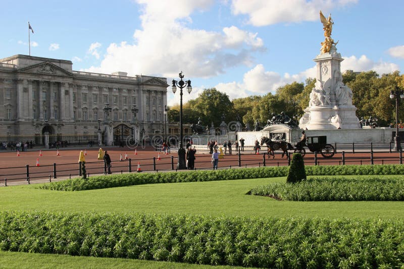 View of Buckingham Palace over green grass and manicured flower bed. No cars. Horse drawn Brougham carriage being ridden in front of the gates. Very few people. Blue sky. Royal Standard flying. View of Buckingham Palace over green grass and manicured flower bed. No cars. Horse drawn Brougham carriage being ridden in front of the gates. Very few people. Blue sky. Royal Standard flying.