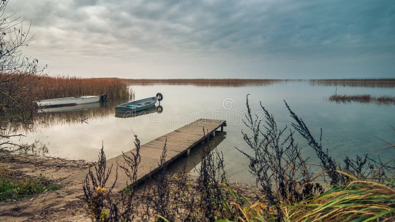 view from the coast of the lake to a beautiful water landscape with a boat, wooden pier and coastal reeds in the light of autumnal stock photos view from the coast of the lake to a beautiful water landscape with a boat, wooden pier and coastal reeds in the light of autumnal stock photos