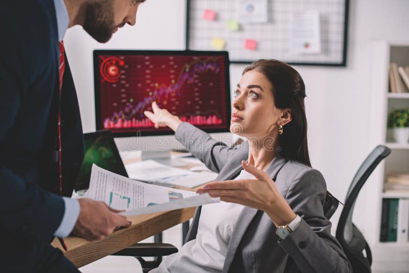 View of data analyst pointing on computer monitor with charts near colleague with papers stock photos