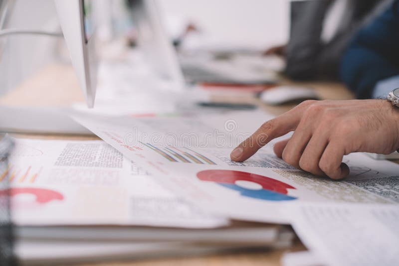 View of data analyst pointing with finger at charts near computer on table stock photo
