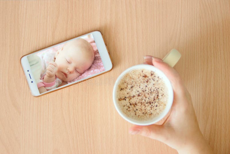 View handheld color video baby monitor. Female hands are holding a smartphone with a baby monitor app. Near hot drink.
