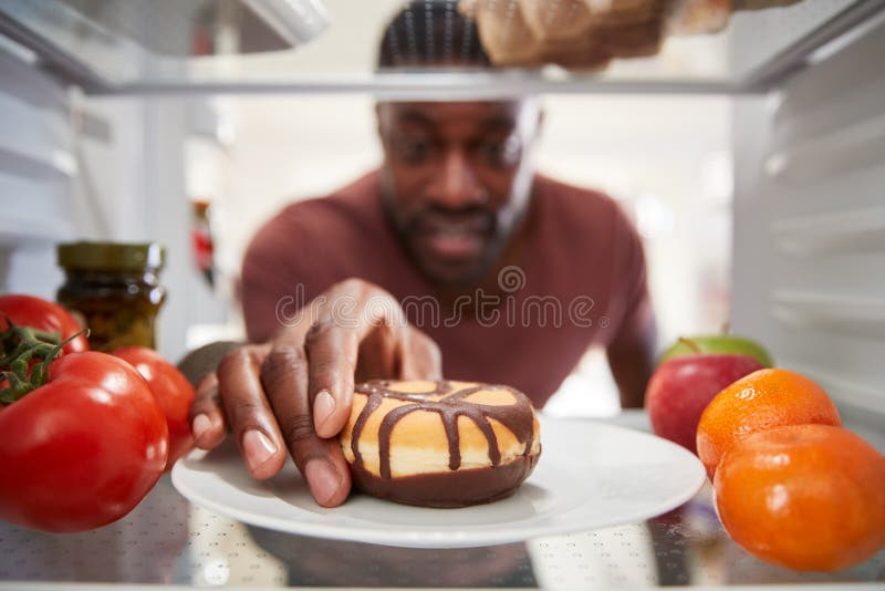 View Looking Out From Inside Of Refrigerator As Man Opens Door And Reaches For Unhealthy Donut stock image