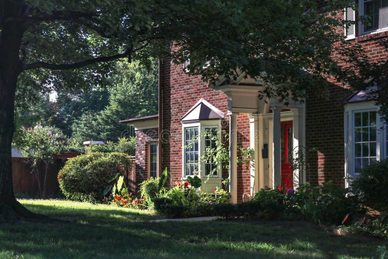 View of pretty two story brick house with bay windows and tall trees and red front door from side angle in late afternoon with hig