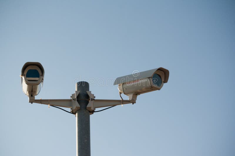 Two security surveillance cameras - one looking directly at the viewer, one directed elsewhere - against a faded blue sky. Two security surveillance cameras - one looking directly at the viewer, one directed elsewhere - against a faded blue sky.