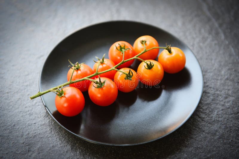 Vino de tomate rojo maduro para la comida cocida en la mesa - Rama de tomates frescos en plato negro con fondo oscuro fotos de archivo libres de regalías Vino de tomate rojo maduro para la comida cocida en la mesa - Rama de tomates frescos en plato negro con fondo oscuro fotos de archivo libres de regalías