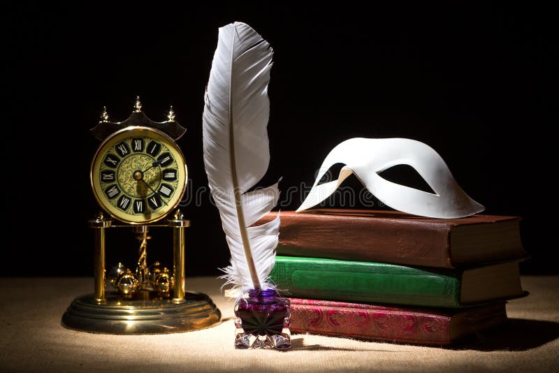 Vintage still life with theater mask on old books near inkstand, feather, old clock against black background under beam of light.