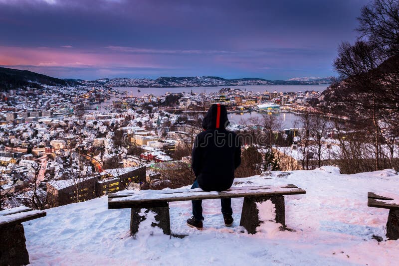 Vinterplats med den ensamma mannen som sitter på den snöig bänken som ser in mot centrum på skymning arkivbild Vinterplats med den ensamma mannen som sitter på den snöig bänken som ser in mot centrum på skymning arkivbild
