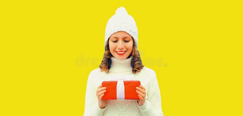 Vinterporträtt av glad leende kvinna som håller en röd presentlåda i händerna med vit trikå som svettare på gul bakgrund