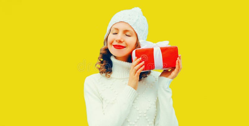 Vinterporträtt av glad leende kvinna som håller en röd presentlåda i händerna med vit trikå som svettare på gul bakgrund
