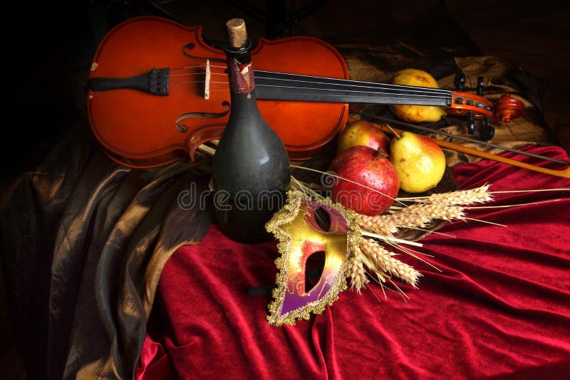 Violino ao lado de uma garrafa do vinho velho e de frutos maduros na tabela, toalha de mesa vermelha de veludo, máscara teatral, foto de stock Violino ao lado de uma garrafa do vinho velho e de frutos maduros na tabela, toalha de mesa vermelha de veludo, máscara teatral, foto de stock