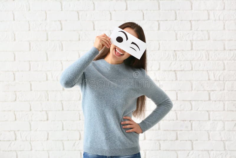 Emotional young woman hiding face behind sheet of paper with drawn eyes against white brick wall. Emotional young woman hiding face behind sheet of paper with drawn eyes against white brick wall