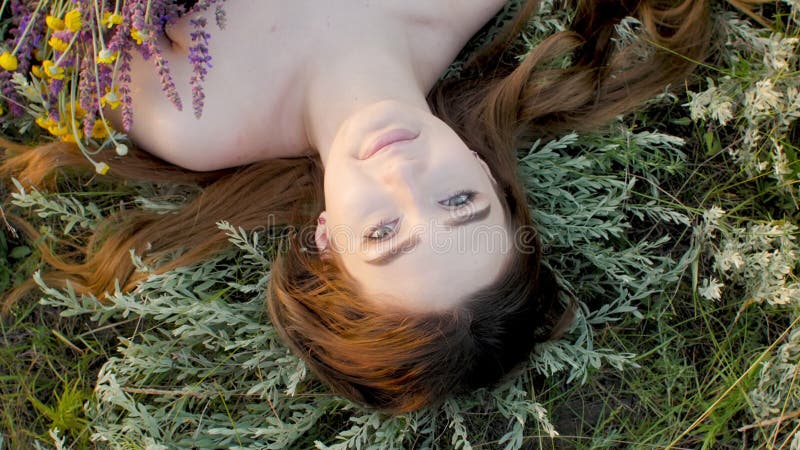 Visage de gros plan de belle fille allongé sur l'herbe avec le bouquet de la jeune femme de fleurs sauvages aux cheveux longs sur banque de vidéos