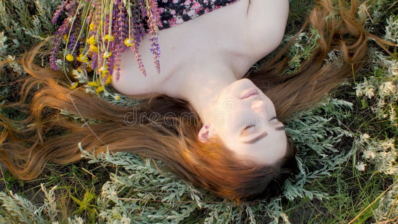 Visage de gros plan de belle fille allongé sur l'herbe avec le bouquet de la jeune femme de fleurs sauvages aux cheveux longs sur banque de vidéos