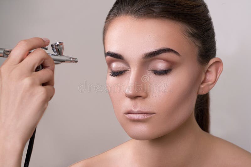 Visagist που κάνει makeup για το πρότυπο με το αερογράφο, που απομονώνεται στο γκρι Υπόβαθρο με το αερογράφο εκμετάλλευσης χεριών στοκ εικόνα με δικαίωμα ελεύθερης χρήσης Visagist που κάνει makeup για το πρότυπο με το αερογράφο, που απομονώνεται στο γκρι Υπόβαθρο με το αερογράφο εκμετάλλευσης χεριών στοκ εικόνα με δικαίωμα ελεύθερης χρήσης