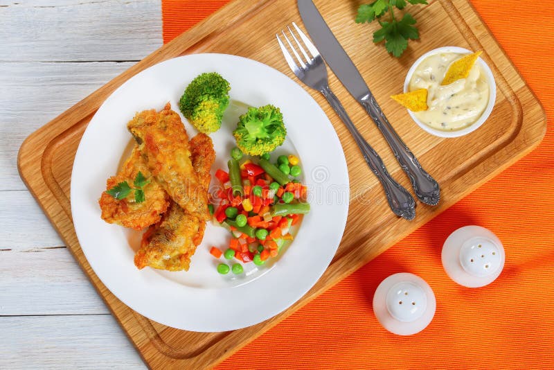 Deep fried perch fillets served with mix of steam cooked vegetables on white plate on chopping board with cutlery and homemade dipping sauce with cornmeal crisps, view from above, close-up. Deep fried perch fillets served with mix of steam cooked vegetables on white plate on chopping board with cutlery and homemade dipping sauce with cornmeal crisps, view from above, close-up