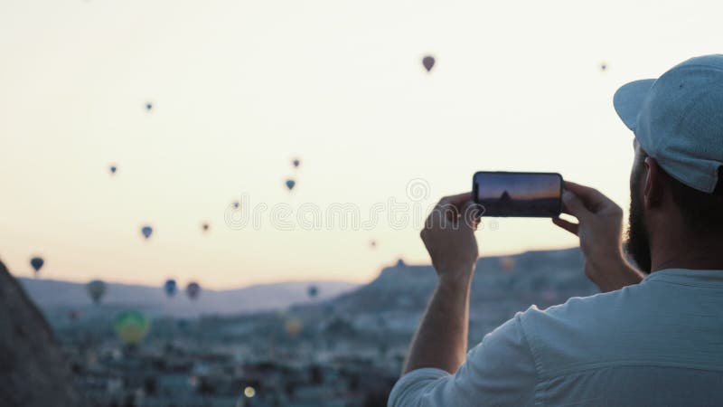 Visión trasera feliz joven turista hombre tomando fotos de increíbles globos de aire caliente con smartphone en cappadocia.