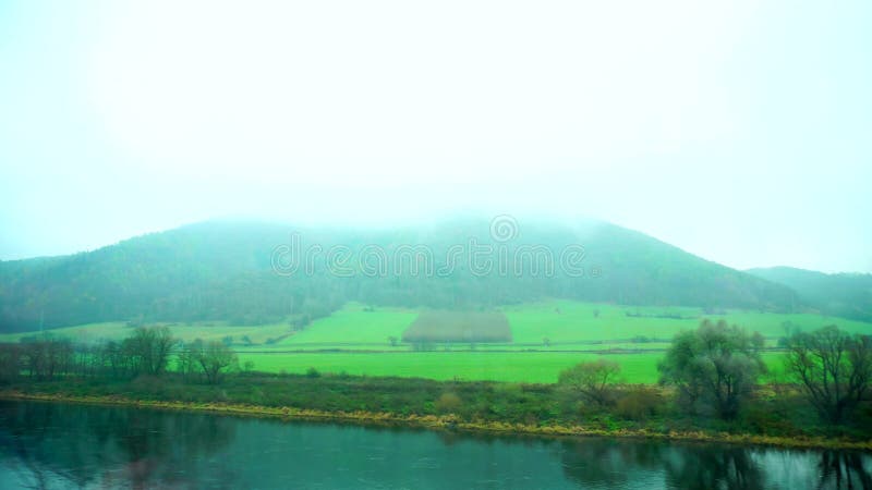 Vista bonita das montanhas na névoa, no rio e em campos verdes da janela de passar o trem no outono