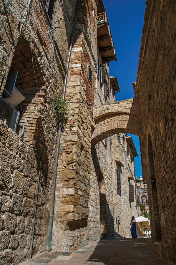 Vista da aleia estreita com as construções, o arco velho e a mulher andando em San Gimignano imagens de stock Vista da aleia estreita com as construções, o arco velho e a mulher andando em San Gimignano imagens de stock