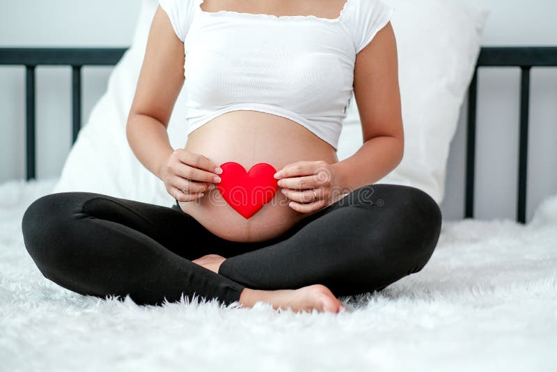 Vista da frente da mulher grávida sentada sobre o leito branco e segurando um símbolo vermelho do coração em sua barriga ou ventr foto de stock Vista da frente da mulher grávida sentada sobre o leito branco e segurando um símbolo vermelho do coração em sua barriga ou ventr foto de stock