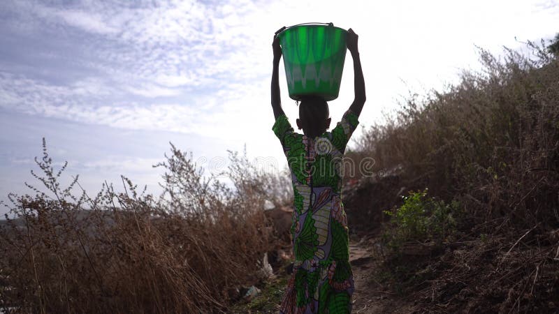 Vista de atrás de una joven africana que llevaba a casa un pesado balde de agua desde un lugar lejos de la aldea almacen de video
