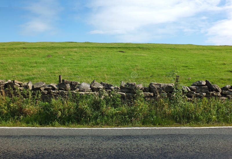 Vista de uma estrada secundária vazia com uma parede de pedra seca coberto de vegetação com uma cerca que separa a de um prado mo foto de stock royalty free Vista de uma estrada secundária vazia com uma parede de pedra seca coberto de vegetação com uma cerca que separa a de um prado mo foto de stock royalty free