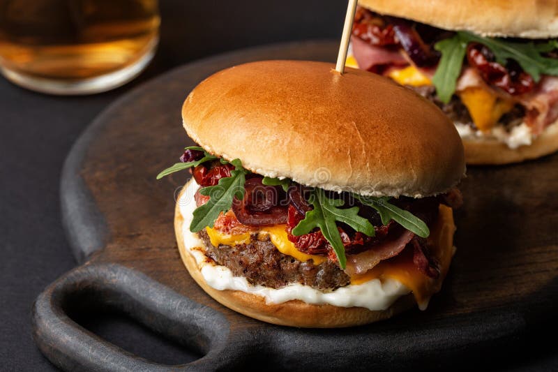 Vista de ángel alto de hamburguesa casera o queso de ternera de hamburguesa frita tocino. Vista de ángel alto de hamburguesa o hamburguesa queso de ternera frito bacon verduras tomates secos al sol y cebollas caramelizadas mayonesa ensalada de cohetes cortada de bollo. vidrio de planta de albahaca de cerveza. fondo de madera