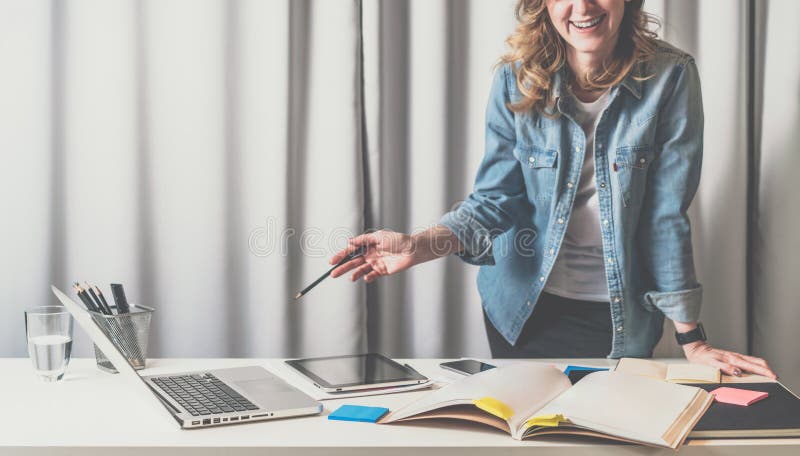 Vista delantera de la mujer joven que se coloca en oficina del diseñador cerca de la tabla en la cual hay catálogos, ordenador po imagen de archivo Vista delantera de la mujer joven que se coloca en oficina del diseñador cerca de la tabla en la cual hay catálogos, ordenador po imagen de archivo