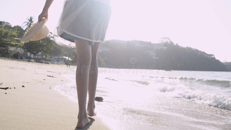 Vista desde la parte trasera de una chica asiática sosteniendo sombrero de paja en un vestido casual y caminando descalzo por la