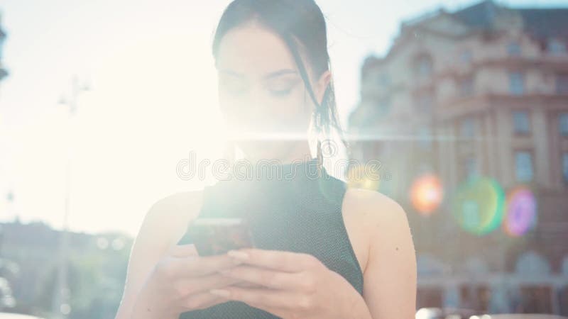 Vista di rotazione di bello castana facendo uso del suo telefono, sorridente felicemente allo schermo, attivamente mandante un sm