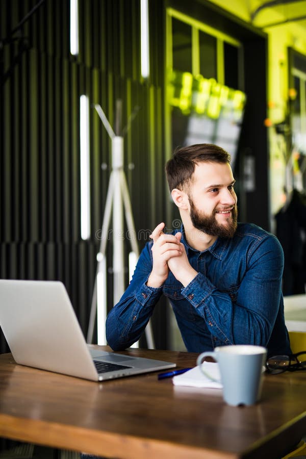 Vista do homem farpado lateral em uma camisa usando um portátil que senta-se em uma tabela em um café na xícara de café da tabela imagem de stock Vista do homem farpado lateral em uma camisa usando um portátil que senta-se em uma tabela em um café na xícara de café da tabela imagem de stock