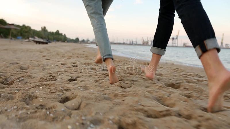 Vista en ángulo bajo, romántica, de dos pies sobre la arena, cogidos de la mano en la playa cerca del mar en cámara lenta. novio