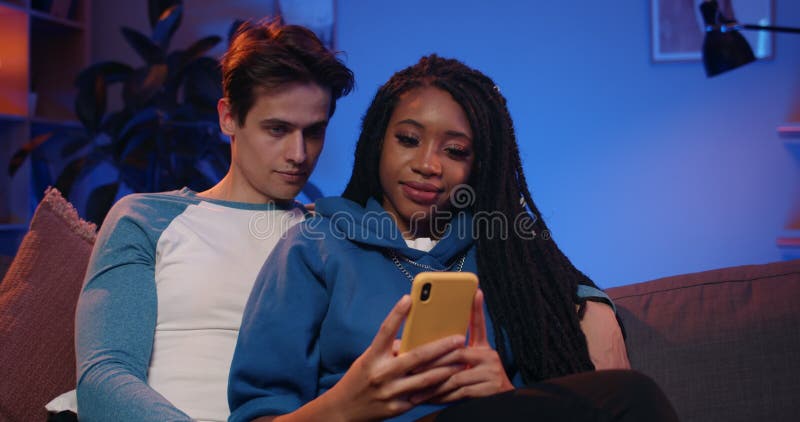 Vista frontal de una joven pareja multiétnica feliz mirando la pantalla del smartphone mientras estaba sentado en el sofá. novio m almacen de video