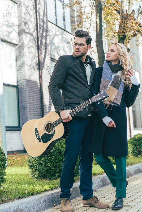 vista integrale di giovani coppie alla moda in attrezzatura di autunno che distoglie lo sguardo mentre stando con la chitarra sul fotografia stock vista integrale di giovani coppie alla moda in attrezzatura di autunno che distoglie lo sguardo mentre stando con la chitarra sul fotografia stock