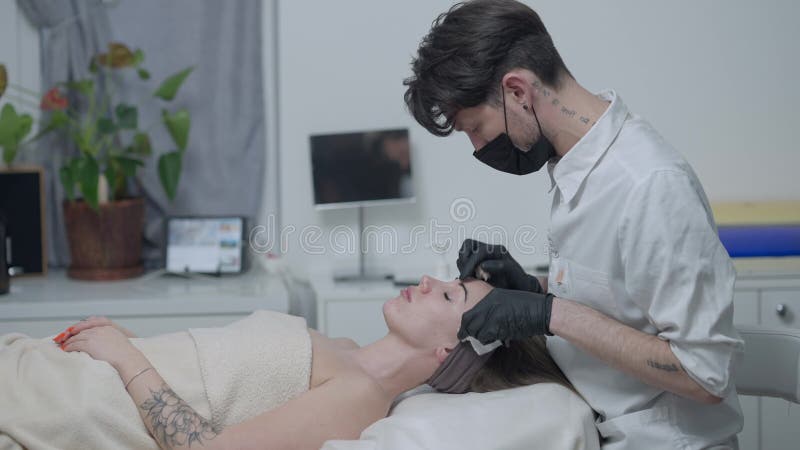 Vista lateral de la esteticista masculina en covid19 máscara facial limpiando la cara de una mujer magnífica en el salón de spa. j