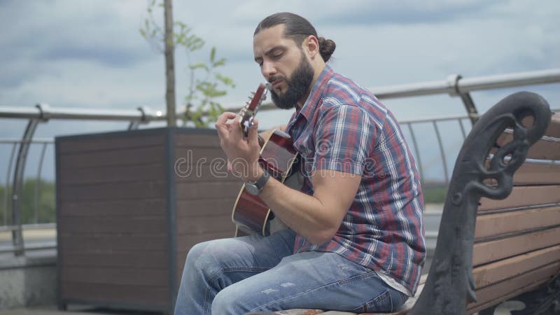 Vista lateral de um guitarrista concentrado sentado no banco urbano no verão Retrato de um jovem caucasiano atencioso