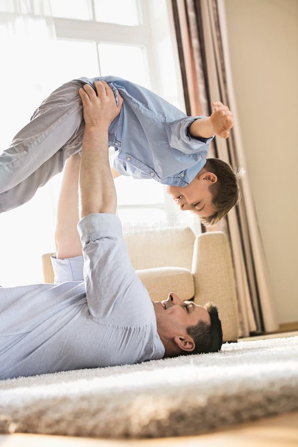 Vista lateral del padre juguetón que coge al hijo mientras que miente en piso en casa fotografía de archivo libre de regalías Vista lateral del padre juguetón que coge al hijo mientras que miente en piso en casa fotografía de archivo libre de regalías