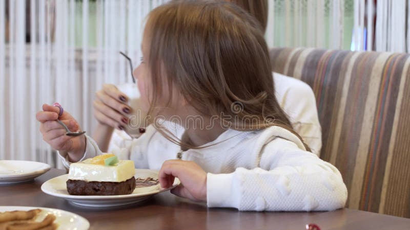 Vista lateral - a menina encantador come a sobremesa que senta-se ao lado da mãe nova bonita