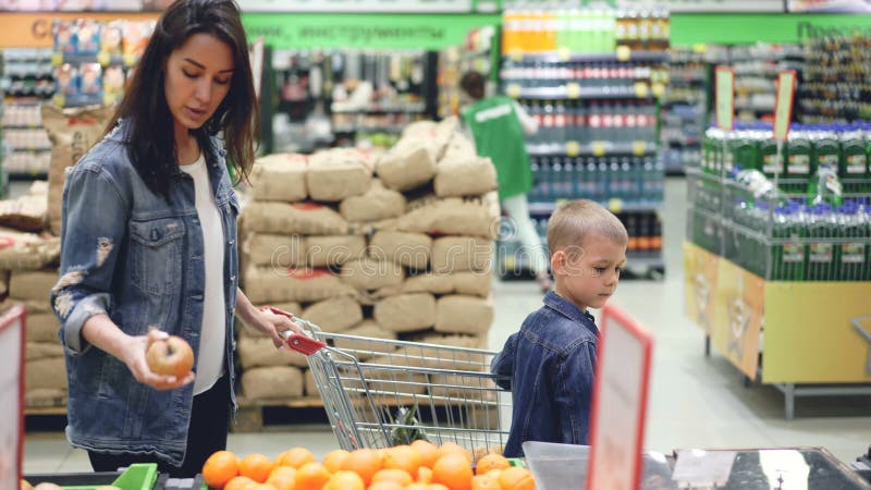 Vista laterale della mamma allegra e del figlio della famiglia che camminano nel supermercato con il carrello di acquisto che sce