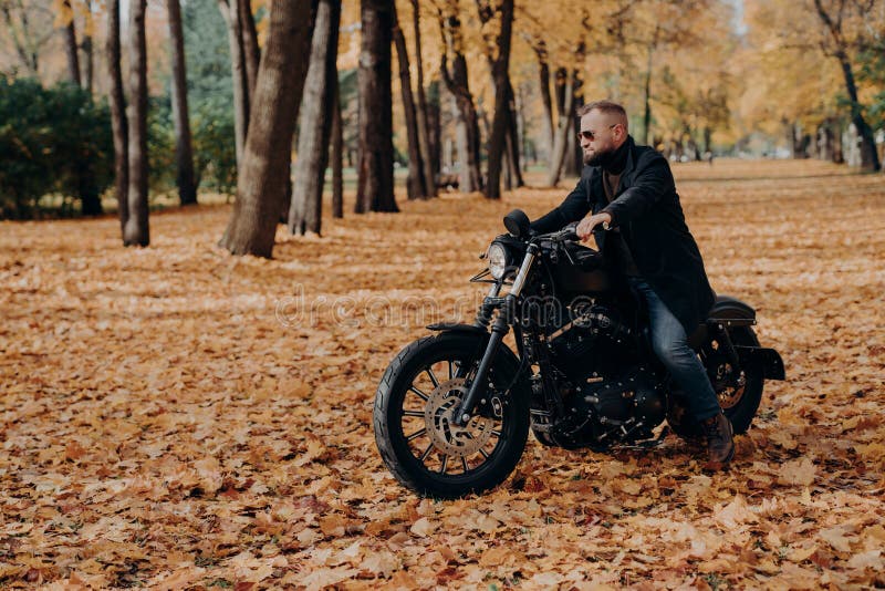 Vista orizzontale all'aperto di motociclisti maschi attivi in bicicletta, con occhiali da sole alla moda e giacca nera, pone in u fotografia stock libera da diritti Vista orizzontale all'aperto di motociclisti maschi attivi in bicicletta, con occhiali da sole alla moda e giacca nera, pone in u fotografia stock libera da diritti