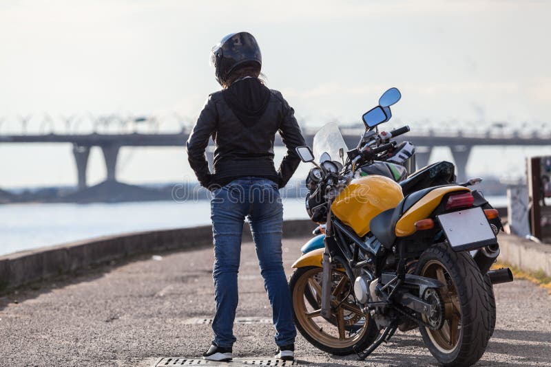 Vista posterior en el motorista de la muchacha en el casco de la motocicleta que se coloca al lado de las bicis en el terraplén d fotografía de archivo Vista posterior en el motorista de la muchacha en el casco de la motocicleta que se coloca al lado de las bicis en el terraplén d fotografía de archivo