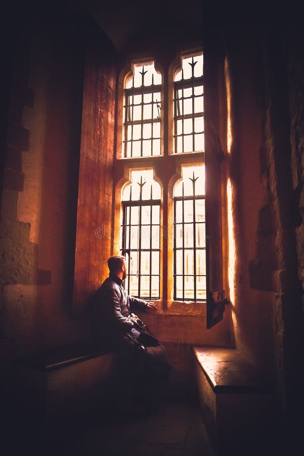 Vista posterior en la silueta del hombre que se sienta en la oscuridad que mira a través de ventana brillante vieja con los rayos fotografía de archivo Vista posterior en la silueta del hombre que se sienta en la oscuridad que mira a través de ventana brillante vieja con los rayos fotografía de archivo