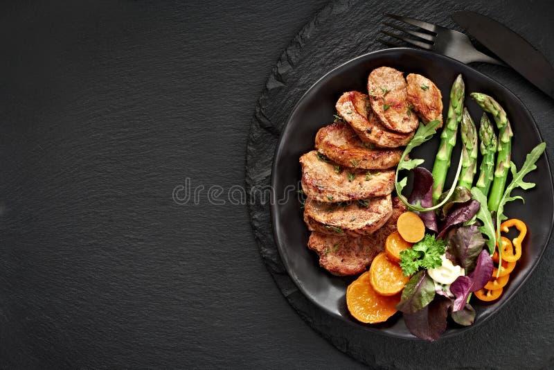Vista superior de un plato negro con rodajas fritas de ternera, batata y ensalada mixta en pizarra negra foto de archivo libre de regalías Vista superior de un plato negro con rodajas fritas de ternera, batata y ensalada mixta en pizarra negra foto de archivo libre de regalías