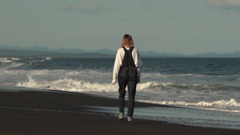 Vista trasera de un modelo irreconocible que camina en una playa de arena contra el fondo de olas que rompen el océano almacen de video