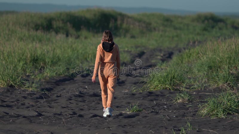 Vista trasera de una mujer irreconocible camina a lo largo de una playa negra con vidrios de hierba verde a hora dorada almacen de video