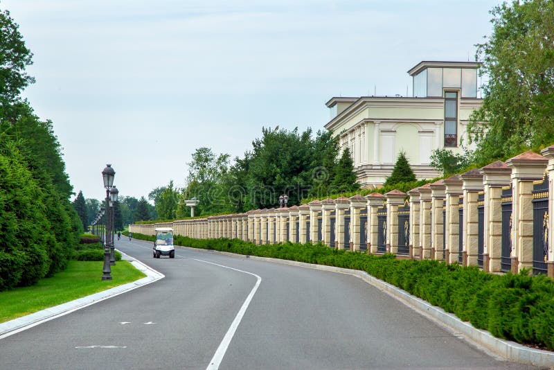 A white golf car rides along an asphalt road along a massive stone fence, cased with green trees growing from bushes and thuja. A white golf car rides along an asphalt road along a massive stone fence, cased with green trees growing from bushes and thuja.