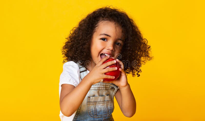 Vitaminsnack. Lilla flicka som biter rött äpple. Lilla flicka som biter det röda äpplet över gul bakgrund.