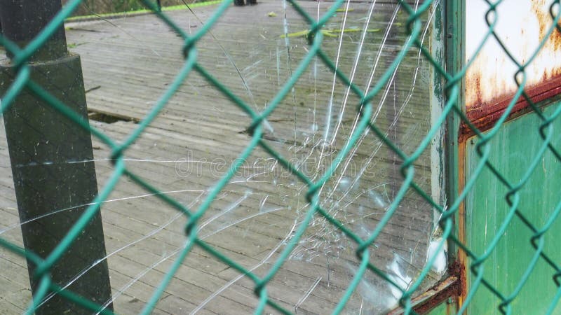 Vitre brisée avec éclats et fissures se rapprochent derrière une clôture maillée de chaîne en flou.